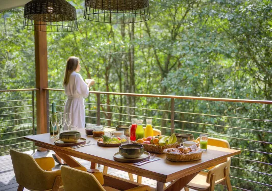 Woman in a white bathrobe looking out to a forest with a table full of food behind her