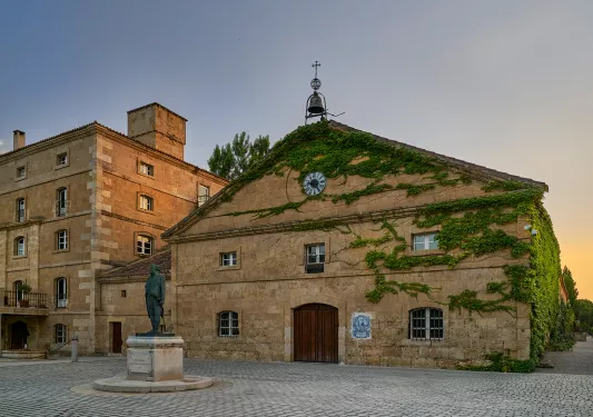 Exterior view of stone building covered in plants, with a bell on top