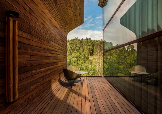 Two wooden chairs and a table in between two buildings looking out to a forest