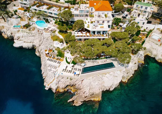 Sky view of a hotel complex with an outdoor pool on a cliff, next to the ocean