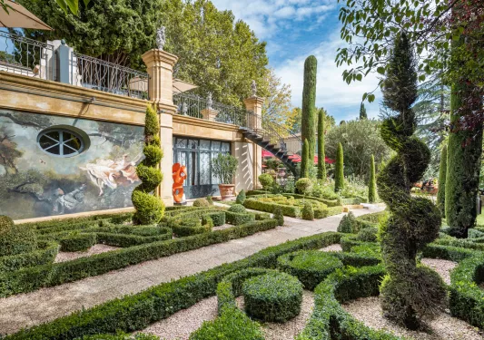 Outdoor garden with trimmed and shaped bushes, with a large building on the left