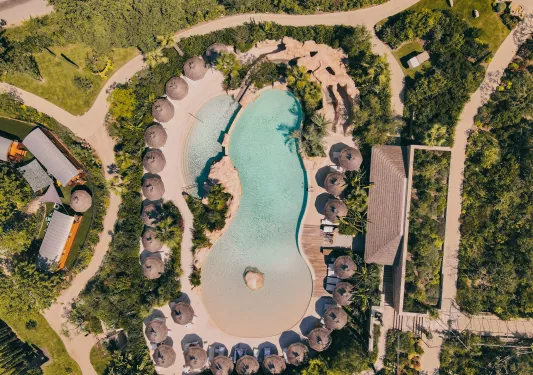Sky view of outdoor pool surrounded by straw umbrellas and trees