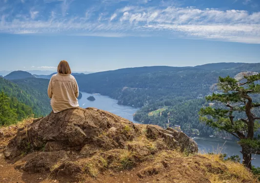 Woman sitting on a large boulder on top of a hill, looking down at the water