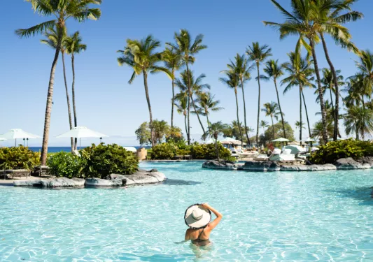 Woman wearing a hat swimming in a pool, surrounded by large palm trees