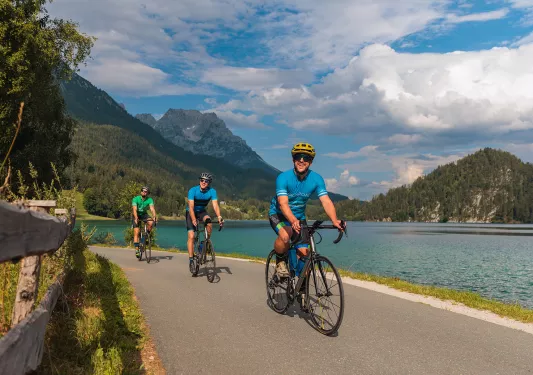 Three men on a road next to a lake riding their bicycles