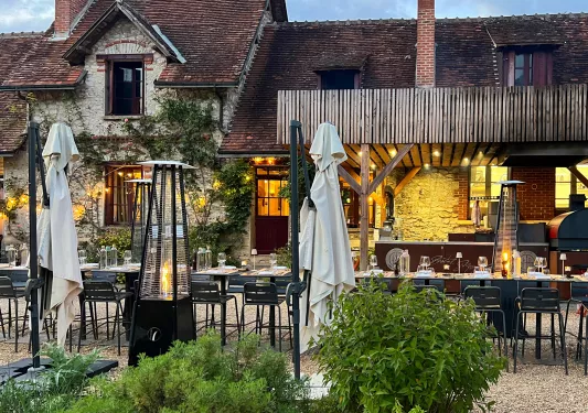 Outdoor dining area in front of a tan and brown brick building, with portable space heaters and umbrellas in the background