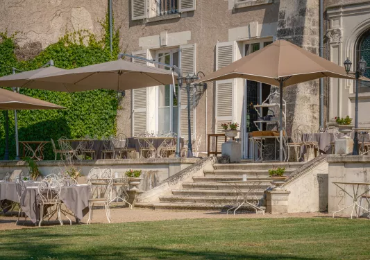 Outdoor patio area with white metal chais, brown umbrellas and a stone building on the right