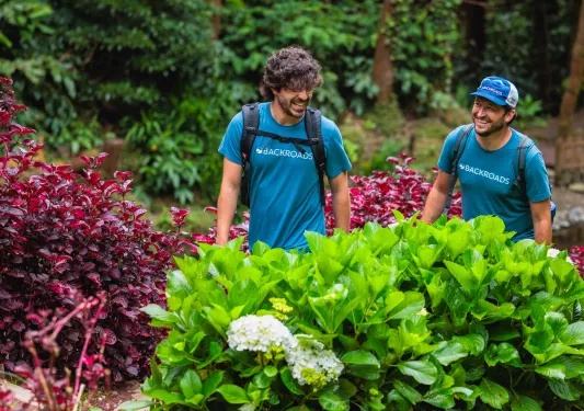 Two men walking through an outdoor path, surrounded by green and red plants