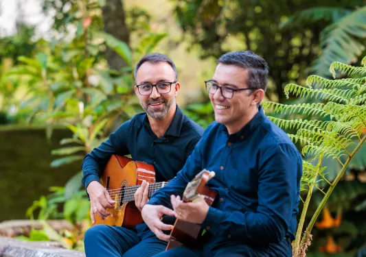 Two men sitting on a stone fence playing guitars and smiling