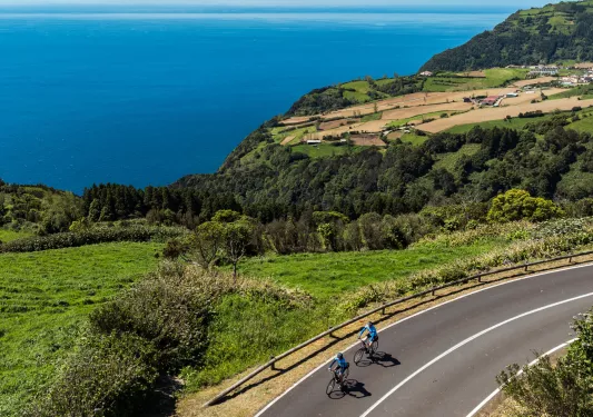 Two people riding their bikes on an asphalt road with a large valley and ocean in the background