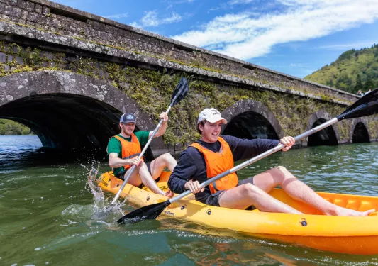 Two men on a kayak, paddling in a river by a bridge