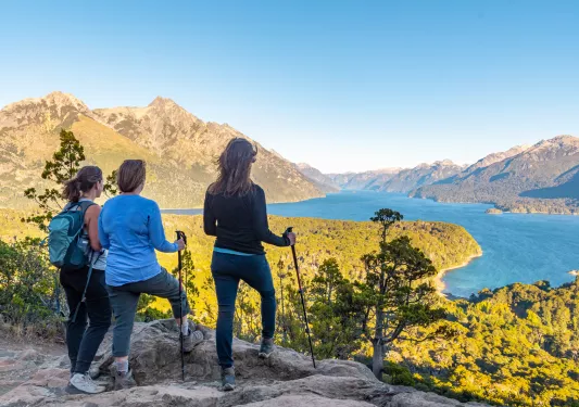 Three women on a cliff, looking out to a valley of trees and a lake