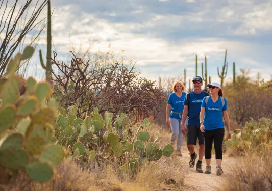Two women and one man hiking on a dirt trail surrounded by cacti and dried trees