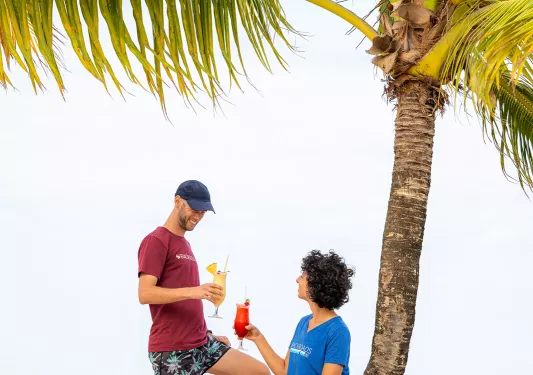 Man and women holding drinks while sitting on a slanted tree on the beach
