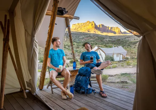Two men sitting on wooden chairs outside of a luxury hut, smiling and laughing