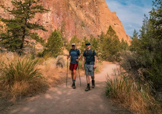 Two men with walking poles, hiking on a dirt trail next to a canyon