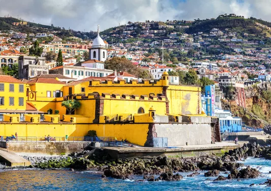 Yellow building by the coast of an ocean, with a hill of houses in the dstance