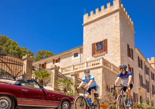 Two men biking and smiling, with a castle-like stone building in the background