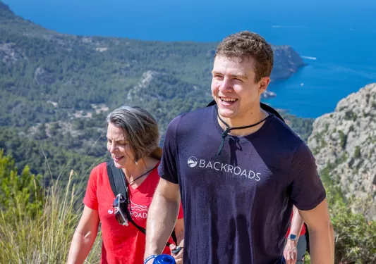 Man and woman smiling while ascending a dirt trail, with the ocean in the distance