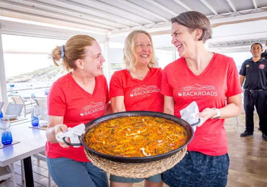 Three women holding up a large pot of cooked food, while smiling at each other