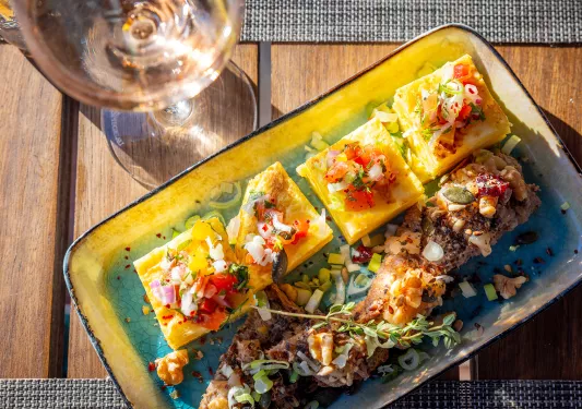A plate full of grilled fish and salad with a wine glass on the left