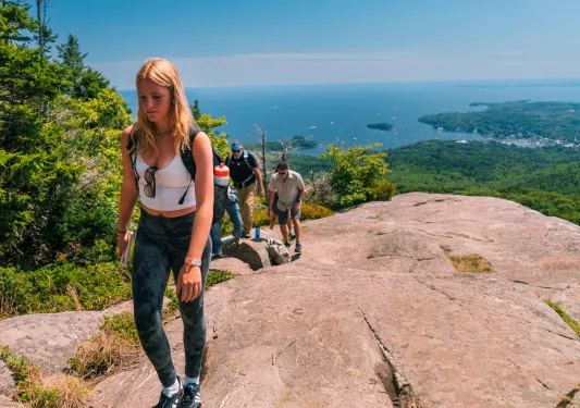 Woman ascending a large boulder on a trail with the ocean in the distance