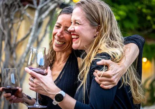 Two women smiling and laughing with arms around their shoulders, holding wine glasses