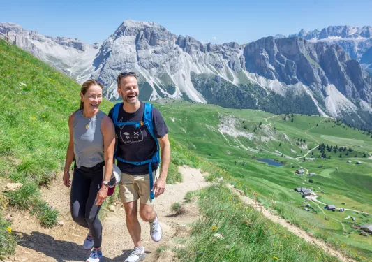 Man and woman smiling, walking on a dirt trail with an open grass valley in the distance