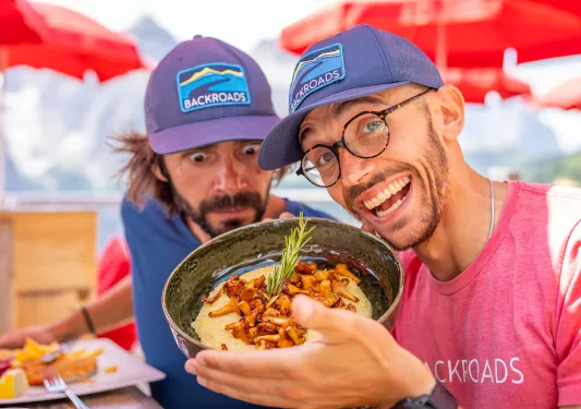 Two men smiling  holding up a bowl of grits and roasted mushrooms