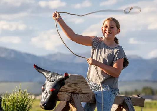 Girl riding a wooden bull prop while swinging a rope