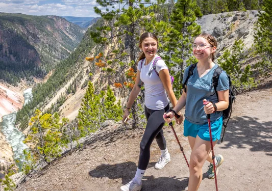 Two girls smiling while hiking, with large mountains and a river in the distance