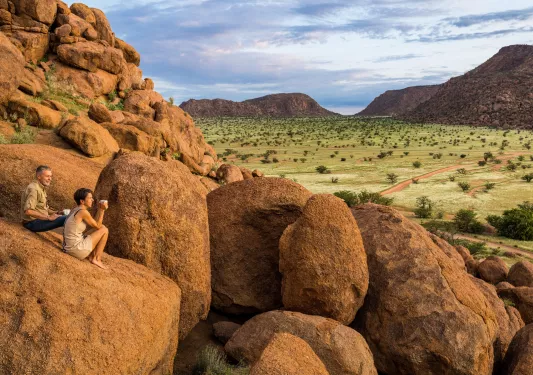 Man and woman sitting on a large boulder overlooking a large, grassy valley