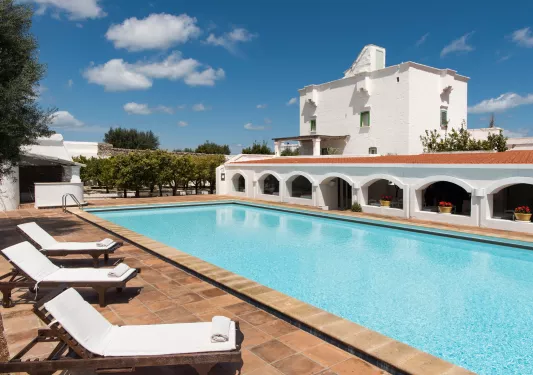 Outdoor pool, surrounded by white pool chairs and a white, rectangular building