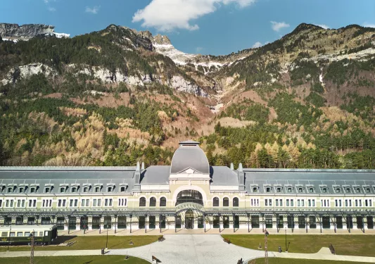 Wide tan and gray building with a large, gray courtyard and tall mountains in the background