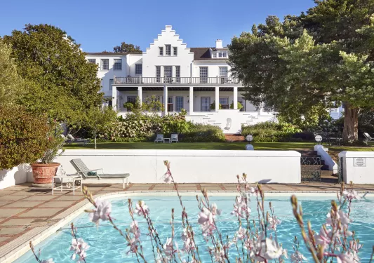Exterior view of white and brown hotel building, with a large grass field and pool in front