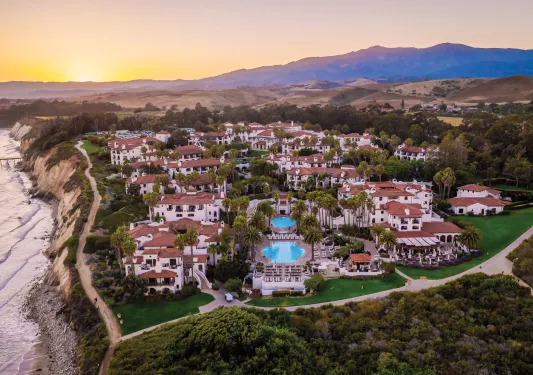 Sky view of white and tan buildings with outdoor pools, with a cliff on the left