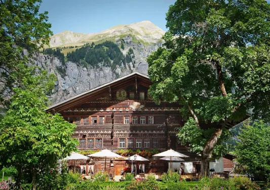 Exterior view of a wooden-style cabin, surrounded by trees and a tall mountain in the background