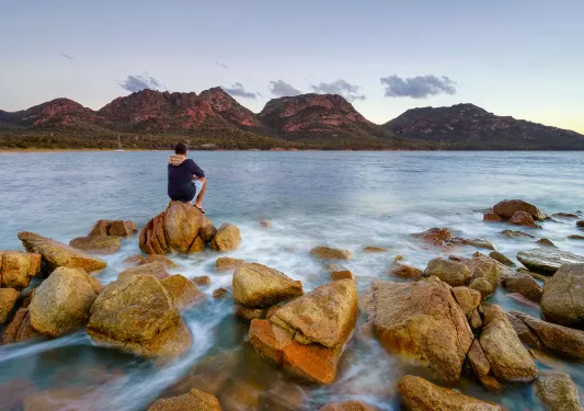 Man sitting on a boulder by the oceanside, looking out to large cliffs