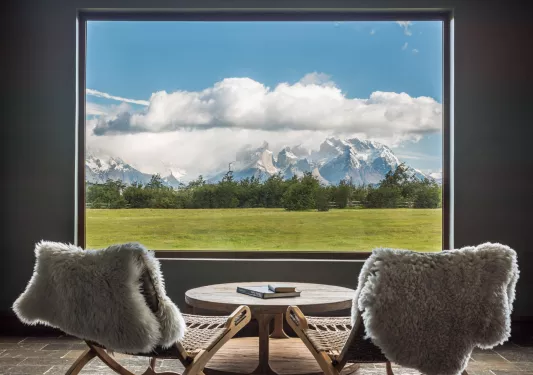 Two woven chairs with fur on them, facing a window looking out to snow capped mountains
