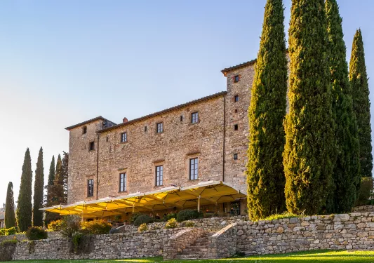 Tall stone building with tall trees and an outdoor patio covered by yellow umbrellas
