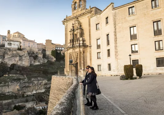 Two women standing on a stone barricade next to a cliff, looking out to other buildings