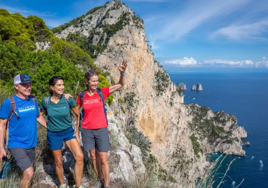 Two women and a man smiling and waving, while standing on a cliff next to the ocean