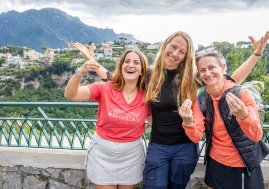 Three women smiling with their arms open, with a town in the background