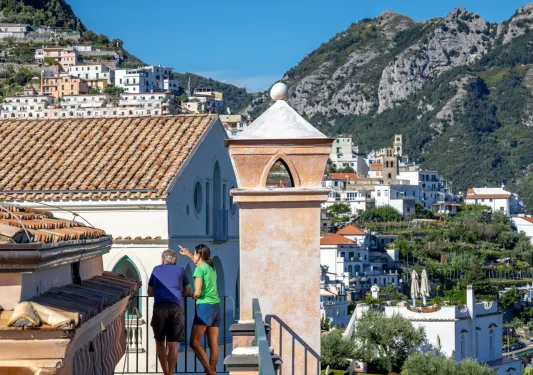 Man and woman on an outdoor balcony, pointing and looking at a town in the distance
