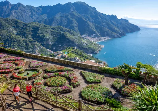Two woman walking through an outdoor garden, with the ocean in the backround
