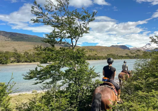 Two women riding horses, going through tall bushes towards a lake
