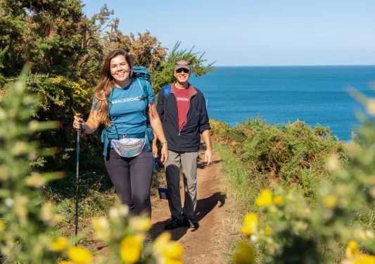 Man and woman with walking poles, ascending a dirt trail with the ocean in the background