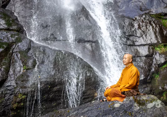 Monk in an orange robe sitting on a large boulder next to a waterfall
