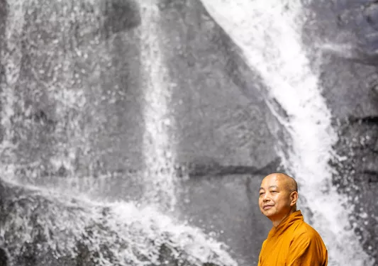 Monk in orange robe sitting on a boulder next to a waterfall