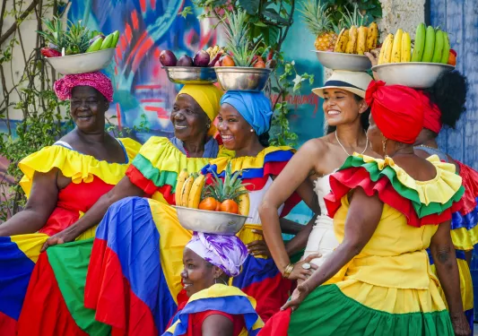 Women in colorful dresses pose with fruit bowls balanced on their heads